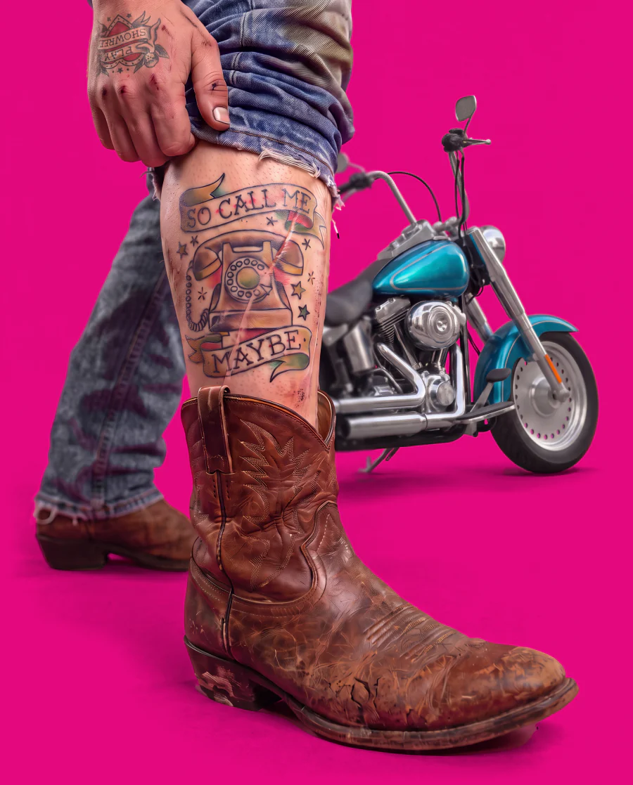 A tattooed hand pulls up a frayed jeans hem over a worn cowboy boot, showing a shin tattoo of a rotary phone that reads “So call me maybe”; a turquoise chopper motorcycle is parked behind.
